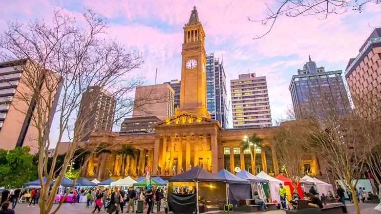 A Brisbane látnivalók közül itt a City Hall óratornya naplementében, a King George Square nyüzsgő közösségi terével és szabadtéri vásárával az előtérben.
