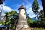 A brisbane-i Old Windmill kőből épült, hengeres történelmi tornya modern felhőkarcolók között, napos városi környezetben.