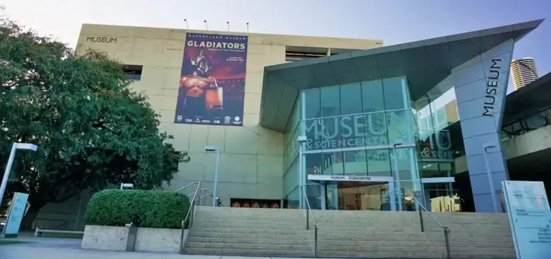 A brisbane látnivalók között a Queensland Museum modern üvegbejárata a MUSEUM felirattal és egy nagyméretű kiállítási bannerrel a homlokzaton.