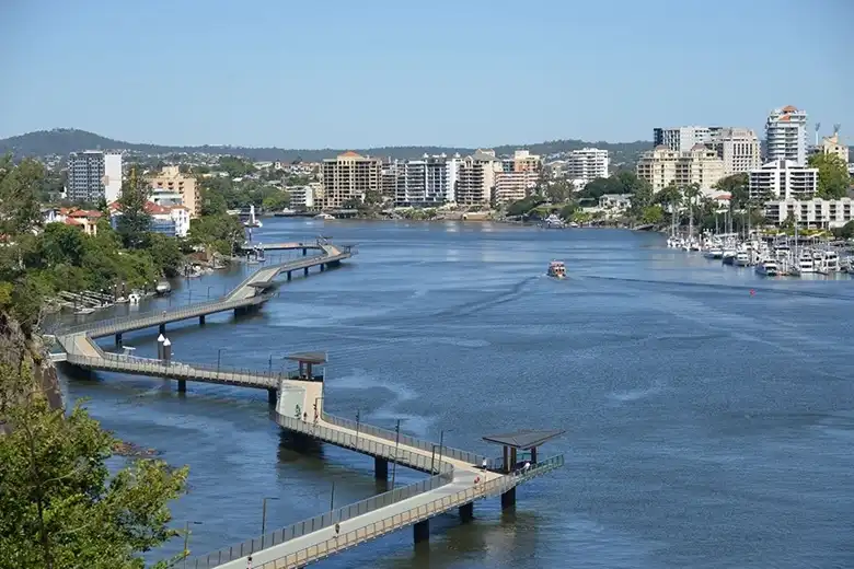 A brisbane-i Riverwalk kanyargó, víz fölé épített sétánya a New Farm felé, modern városi háttérrel, kikötővel és napfényes folyóparti panorámával.