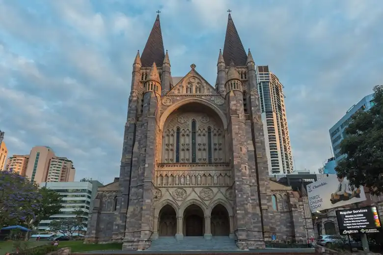 A brisbane-i St John’s Cathedral gótikus homlokzata három toronnyal, modern felhőkarcolók között, napfényes városi környezetben.