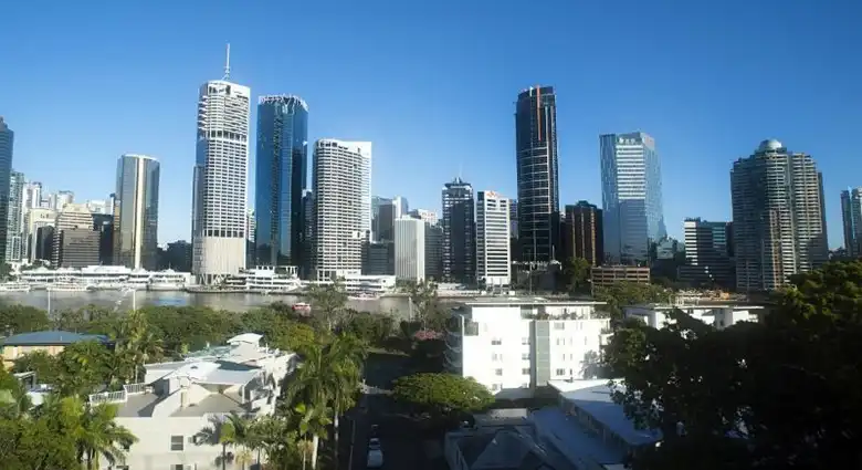 Brisbane modern felhőkarcolói a folyópart fölé emelkedve, előtérben zöld lakóövezettel és tiszta, napos városi panorámával.