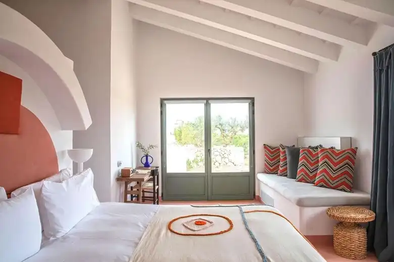 Mediterrán stílusú hotel szoba Menorcán, fehér ágy, terrakotta íves fejtámla, mintás párnák és nagy üvegajtó kertre néző kilátással.